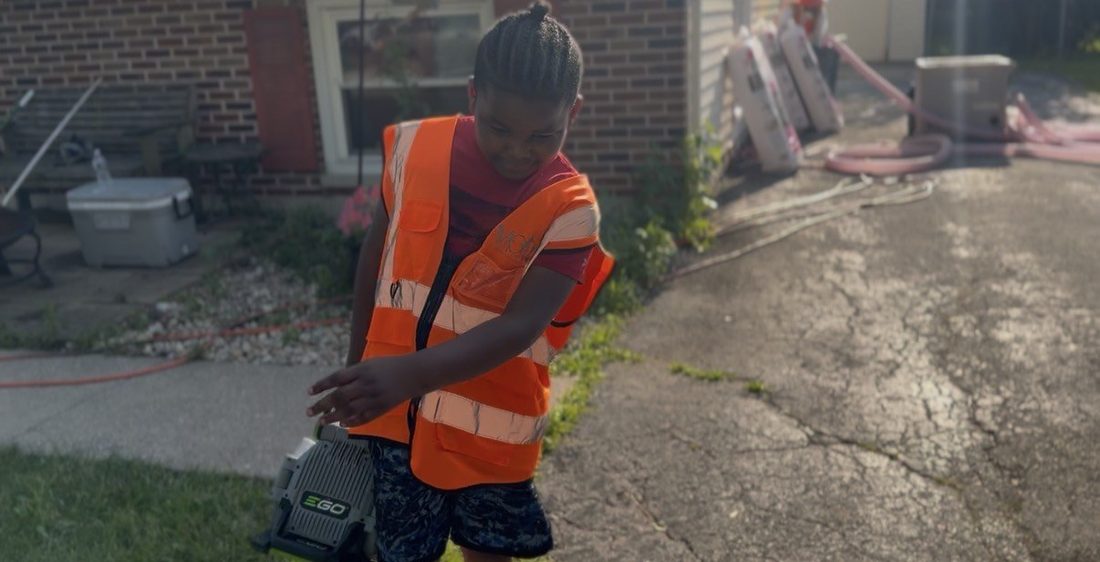 A young V.O.V helper in a safety vest carrying a battery blower